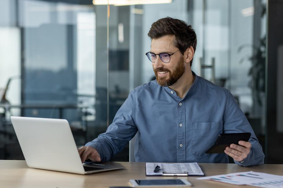 Lächelnder junger Finanzexperte und Steuerberater arbeitet im Büro am Laptop bei Pandotax mit Dokumenten und Taschenrechner. Er kalkuliert die monatlichen Umsatzsteuervoranmeldungen.